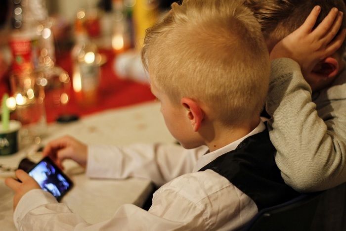 Ilustrasi dua bocah lelaki sedang menonton sesuatu dari layar smartphone saat duduk di meja makan.