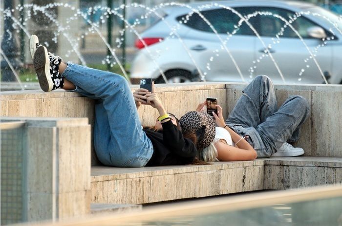 Ilustrasi dua remaja putri memainkan ponsel mereka dengan tiduran di atas bangku taman kota.